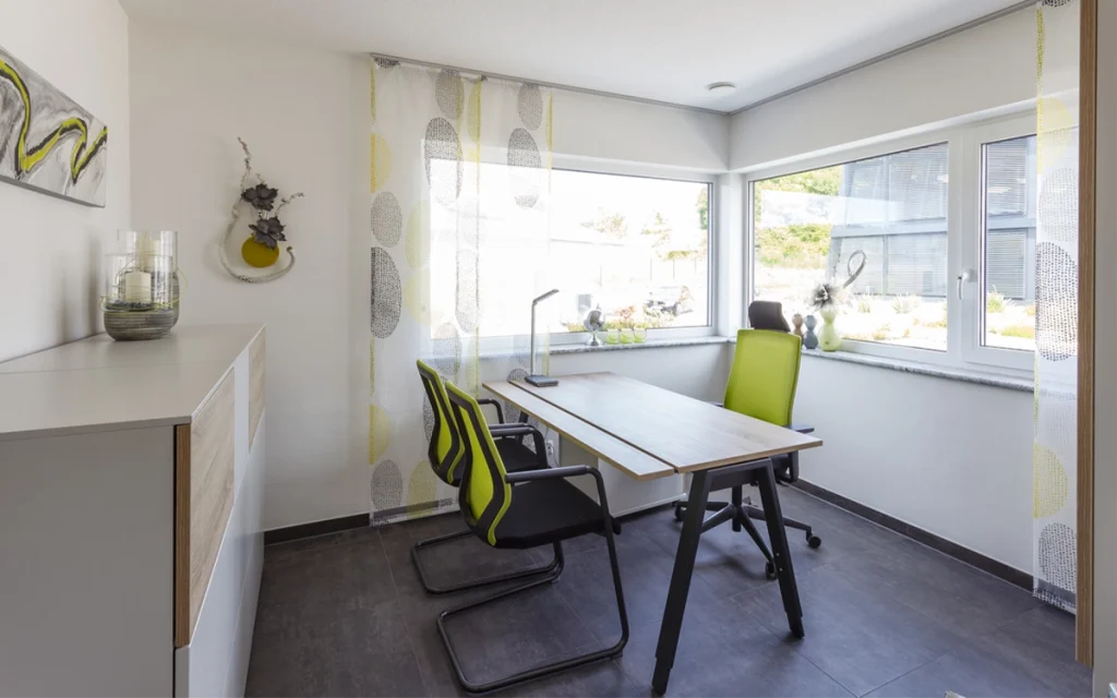 Modern eingerichtetes Büro mit Schreibtisch und Stühlen vor Fenstern.