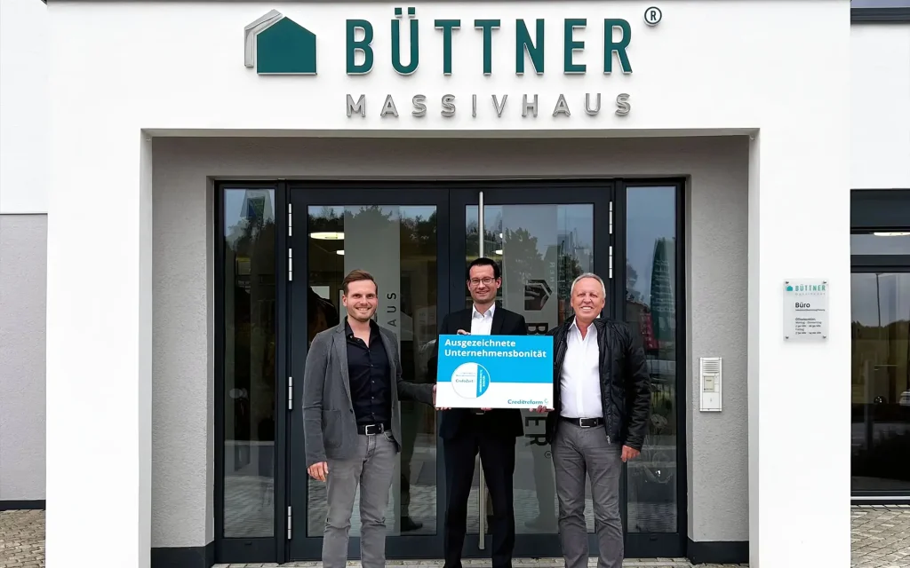 Drei Personen vor dem Eingang von Büttner Massivhaus mit einer Auszeichnung.