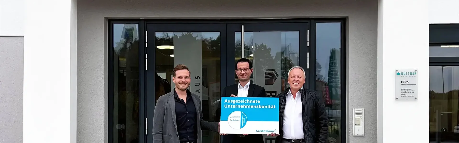 Drei Personen vor dem Eingang von Büttner Massivhaus mit einer Auszeichnung.