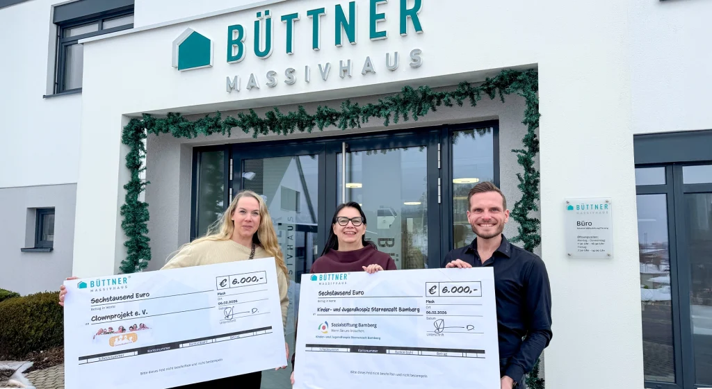 „Drei Personen stehen vor dem Gebäude von Büttner Massivhaus und halten zwei große Spendenschecks über jeweils 6.000 Euro für gemeinnützige Organisationen in die Kamera.