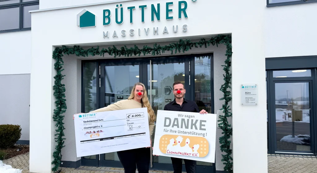 ZweiPersonen stehen vor dem Gebäude von Büttner Massivhaus und halten einen großen Spendenscheck über 6.000 Euro in die Kamera.