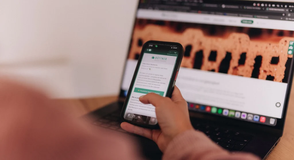 Person nutzt Smartphone und Laptop, um digitale Inhalte und Newsletter von Büttner Massivhaus aufzurufen.