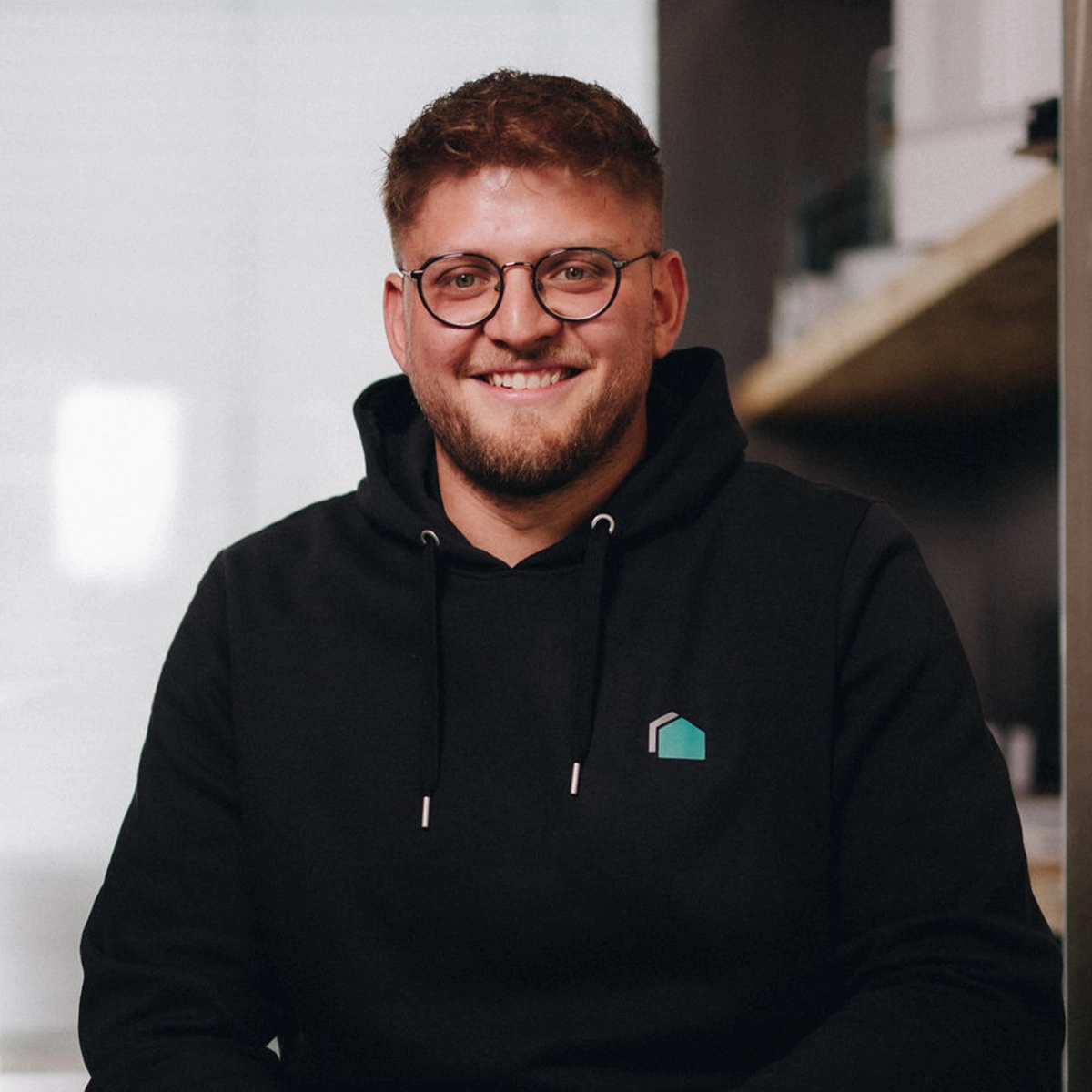Erik lächelt in die Kamera und trägt einen schwarzen Hoodie mit Büttner-Logo sowie eine runde Brille.