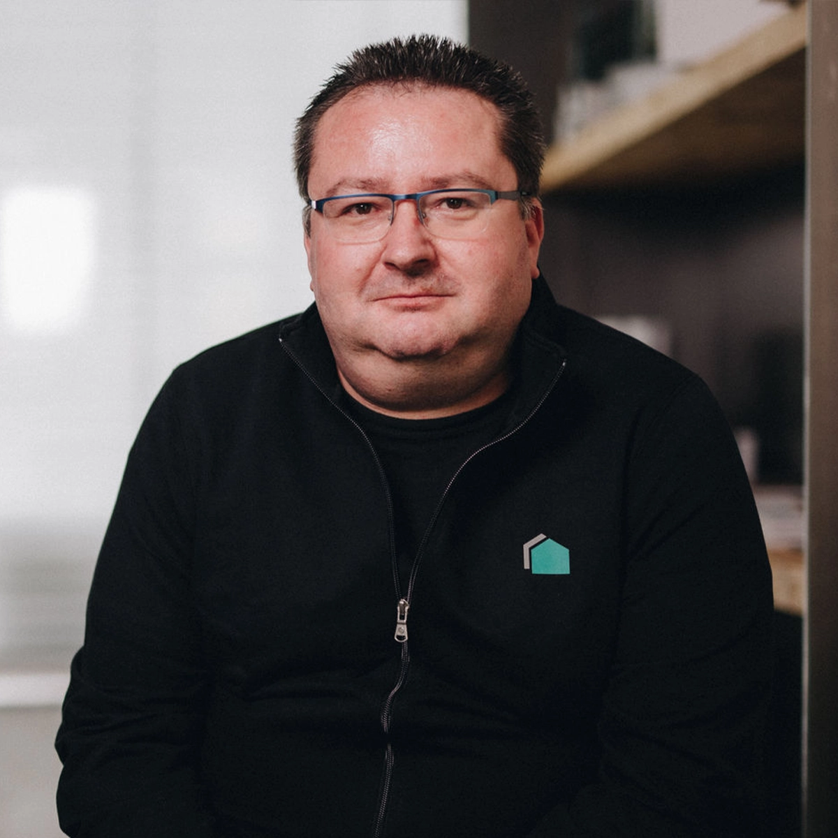 Martin sitzt vor Regalen und trägt eine schwarze Jacke mit Büttner-Logo sowie eine Brille.