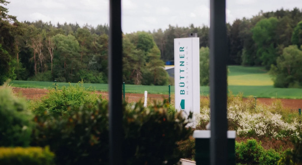 Blick auf das Büttner Massivhaus Gelände mit Stele vor einer idyllischen Naturkulisse aus Wiesen und Wald.