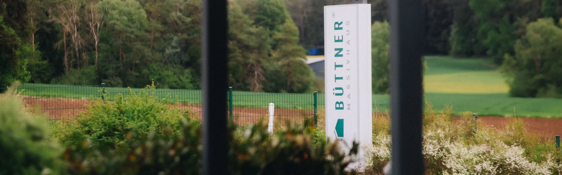 Blick auf das Büttner Massivhaus Gelände mit Stele vor einer idyllischen Naturkulisse aus Wiesen und Wald.
