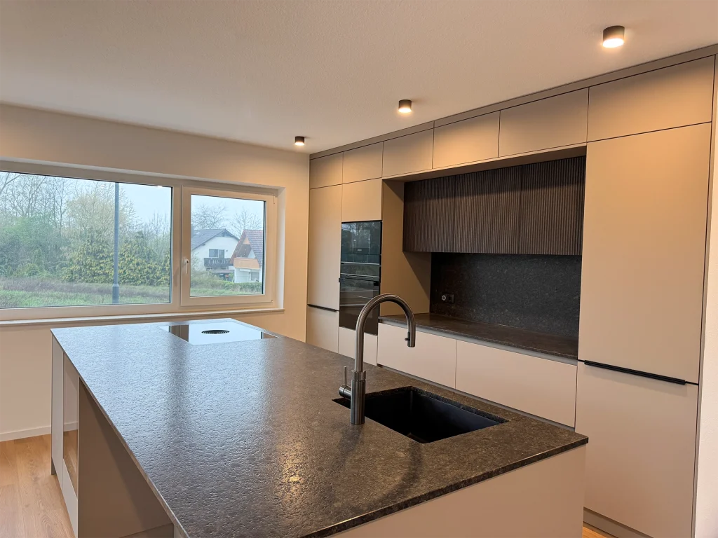 Moderne Küche mit großzügiger Kochinsel, integrierten Geräten und großem Fenster für viel Tageslicht.